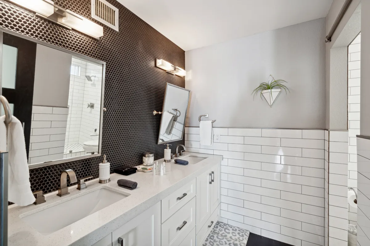 A modern bathroom featuring a double vanity with white cabinetry, expansive mirrors, a decorative plant, and a dramatic black hexagonal tile accent wall. The space feels both functional and spa-like—perfect for morning routines or evening wind-downs.