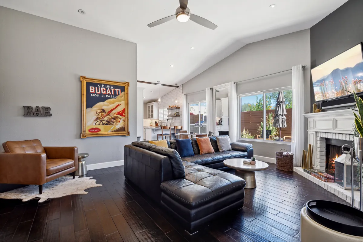 A spacious, contemporary living area featuring a large black sectional sofa, striking vintage Bugatti poster, modern fireplace, a stylish bar area, and expansive windows that flood the space with natural light. The room is arranged for group gatherings, with plenty of seating and an inviting, open layout.