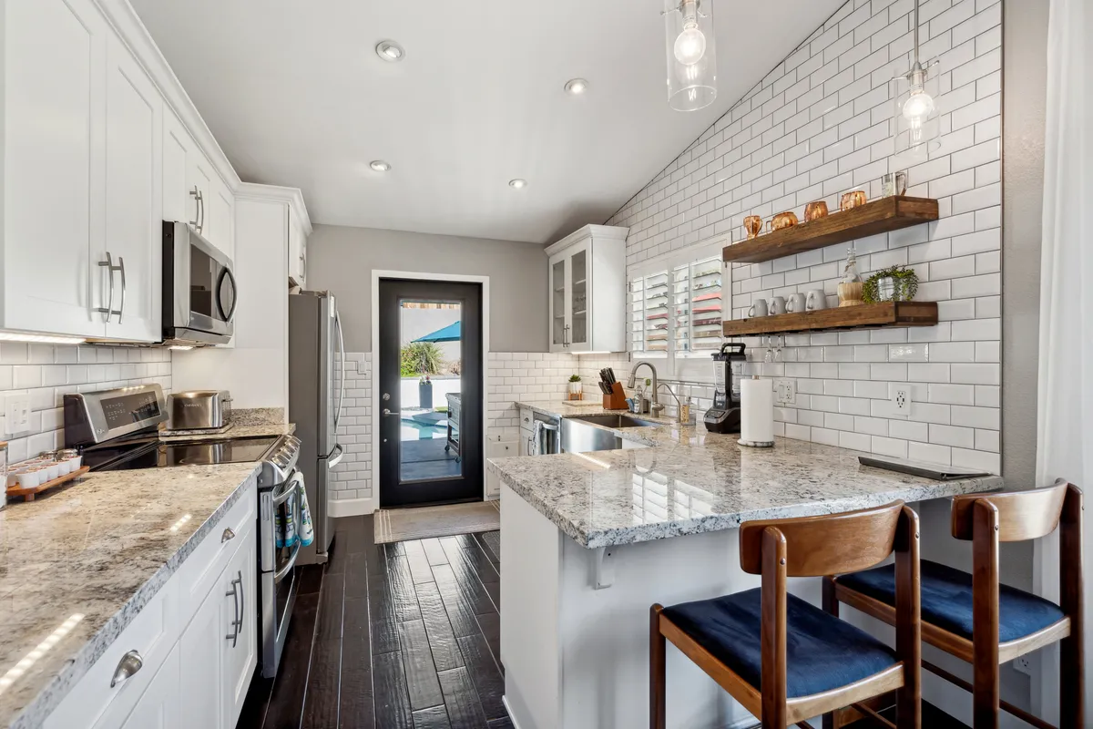 A modern kitchen with granite countertops, open shelving, and gleaming stainless steel appliances. The kitchen opens to the living space and includes a black door leading outside, suggesting a seamless flow for entertaining and group activities.