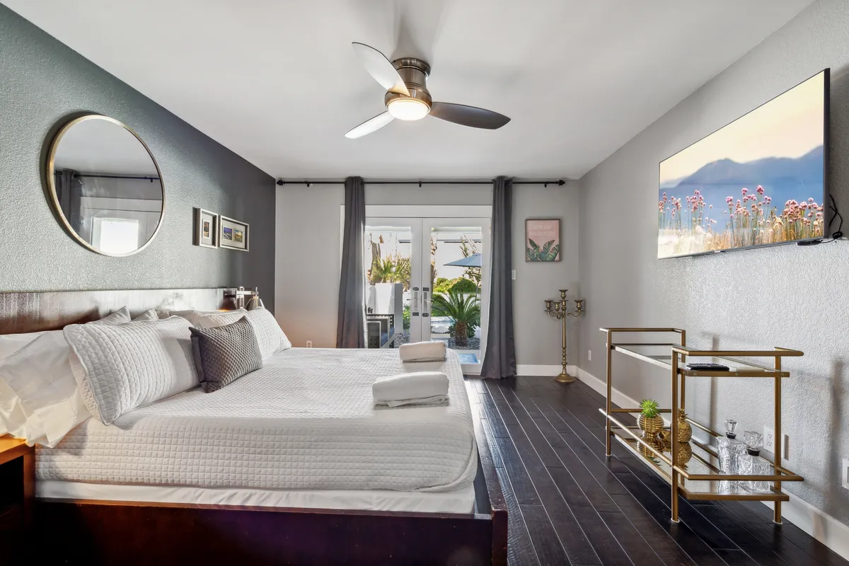 A serene primary bedroom with a California King bed dressed in white linens, a circular mirror above the headboard, sliding glass doors opening directly to the pool area, a wall-mounted smart TV, and a gold accent table. Natural light streams in, enhancing the restful atmosphere.