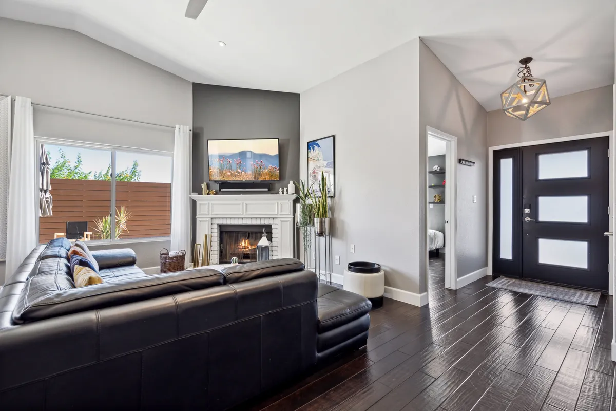 This image showcases a welcoming living area with a fireplace, large windows, a leather sofa, a TV, and a visible entrance door. The room feels cozy and secure, with natural light and a view of the entryway, conveying a sense of comfort and a safe environment.
