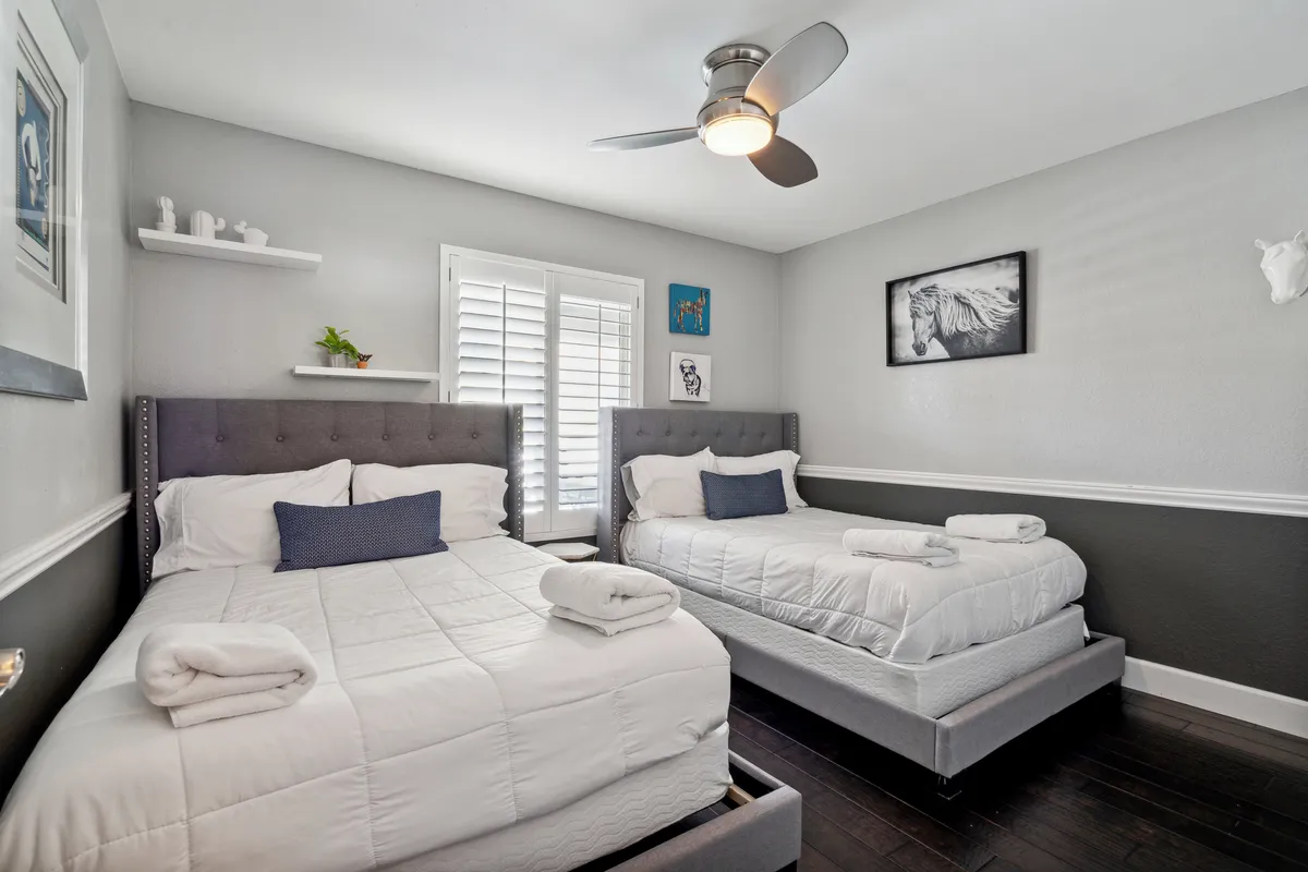 A guest bedroom featuring two Full beds with gray upholstered headboards, elegant wall art, and natural light filtered through window shutters. The space is airy, cozy, and welcoming—ideal for family or friends traveling together.