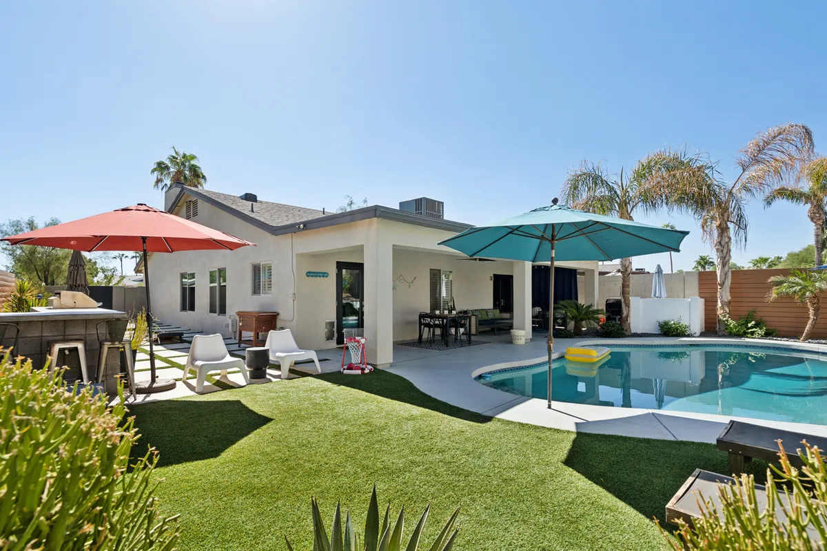 The image showcases a sparkling heated swimming pool at sunset, surrounded by lush green grass, vibrant umbrellas, and plenty of inviting outdoor seating. The cheerful scene hints at fun poolside gatherings, relaxed sunbathing, and moments of leisure under the Arizona sky.