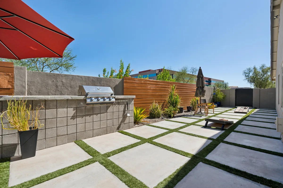 A casual, welcoming patio scene featuring a grill, outdoor seating, umbrella, and paved pathways. The setup invites guests to gather for barbecues, alfresco dining, and celebrations, highlighting the versatility of the backyard.