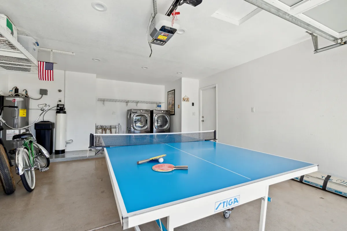 An interior space with a ping pong table, washer and bicycle visible in the background. The area conveys a playful, relaxed mood, suggesting opportunities for friendly competition and casual fun.