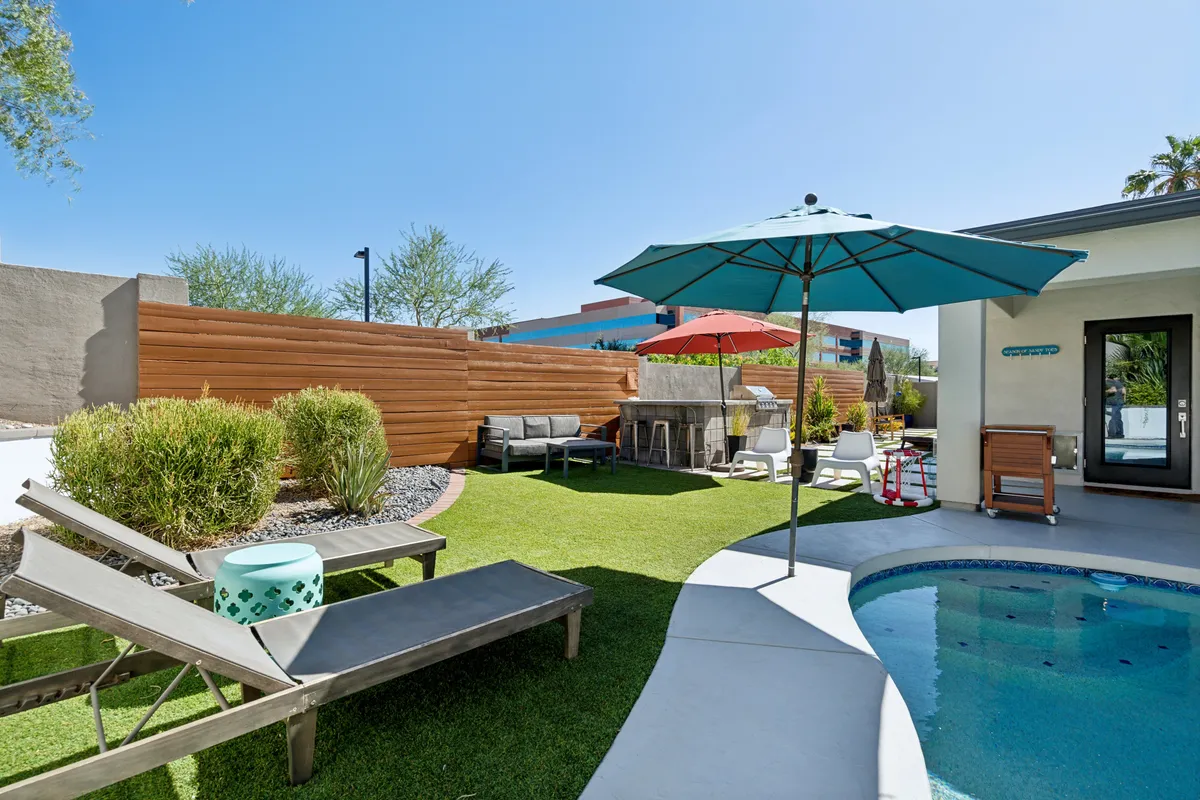 A sunlit backyard featuring a sparkling swimming pool surrounded by lounge chairs, umbrellas, a barbecue area, and a green lawn—perfect for outdoor games and family gatherings. The overall mood is lively and inviting, highlighting amenities for both relaxation and active play.
